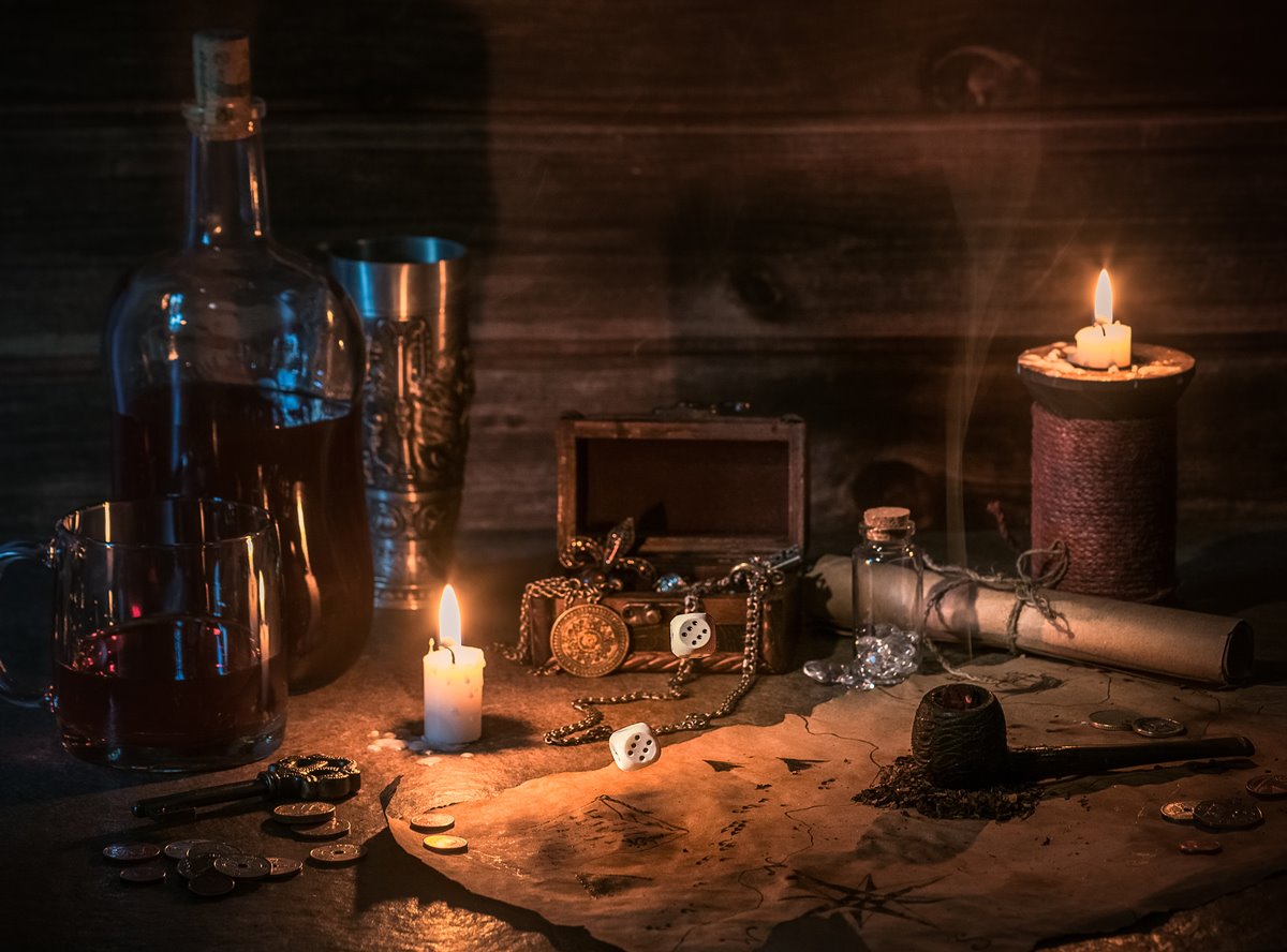 article thumb - Candles light a table. On the table is a map, coins, dice, a smoking pipe and a bottle of rum 