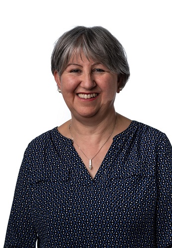 Angela is a woman with short grey hair. She is wearing a dark blue patterned top and a silver necklace
