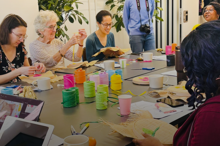 A group of people sitting around a table participating in a raffia embroidery workshop. Colorful spools of raffia thread, cups, and craft materials are spread across the table. Some participants are stitching on the raffia, while others smile and chat. A woman standing at the back, wearing a blue sweater and camera around her neck, laughs with the group. Large green plants decorate the bright, welcoming room.
