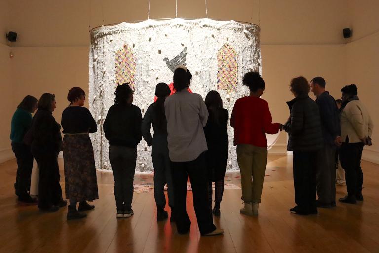 People stand in a dark art gallery. They are gathered around a large installation that is spot lit. The installation is an alter-type structure made of beads. 