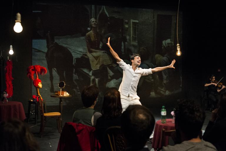A theatrical performer in white gestures expressively on stage in front of a seated audience.