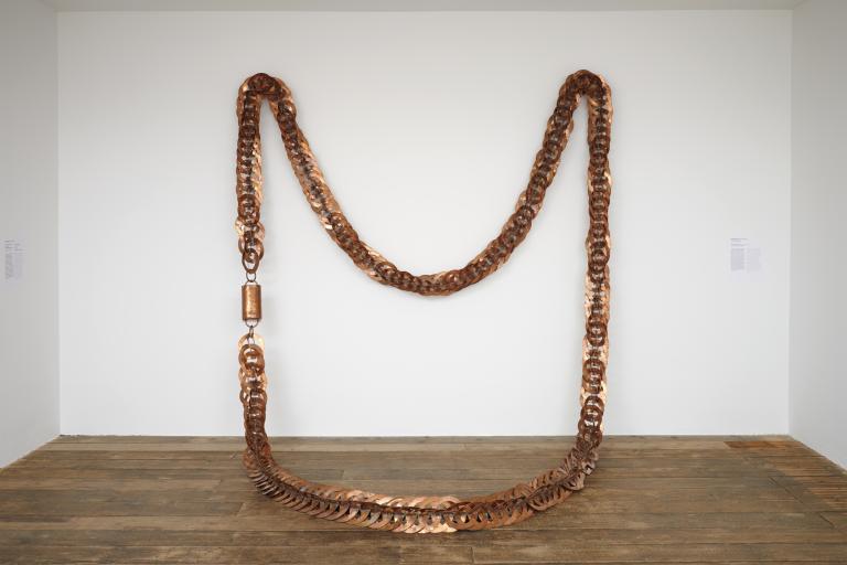 A large sculpture hanging in an art gallery. The sculpture is a giant copper necklace. 
