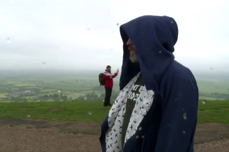 An image of Wes Viola in a dark blue hooded robe. In the back ground are the fields of Somerset, and a man wearing orange taking a picture, who due to an illusion of perspective appears much smaller than the figure of Wes in the foreground