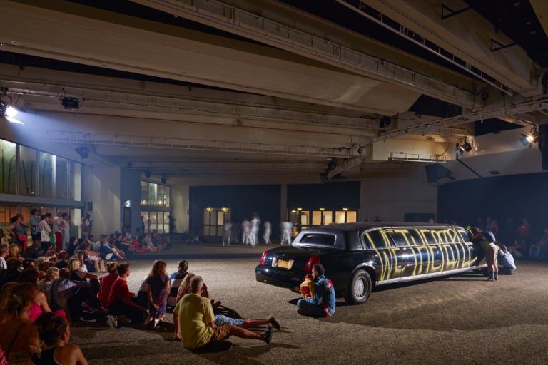 Audiences sit in front of a black limousine in a concrete space.