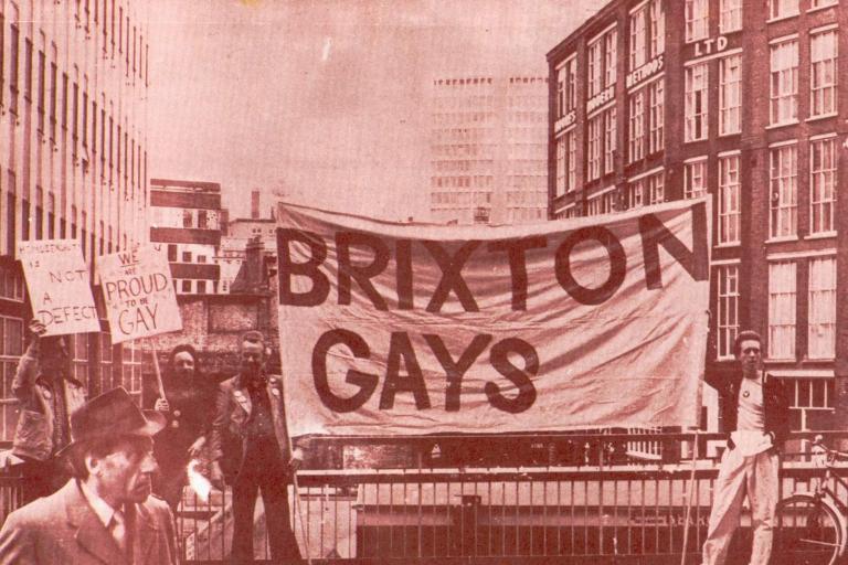 A sepia tone image of a protest march from the 1970s, with two men holding up a large banner that reads 'Brixton Gays'
