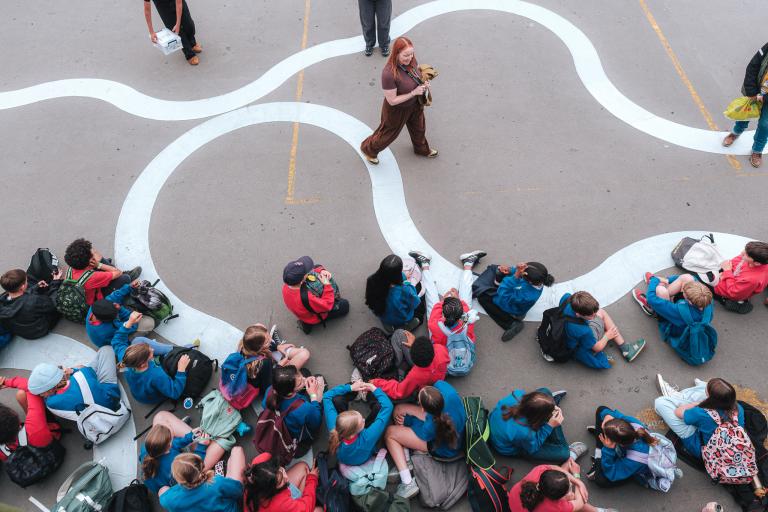 A birdseye view of a group of schoolchildren sitting down on the floor being spoken to by a facilitator who is standing in front of them