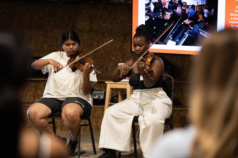 Photo of two people sitting down, playing the violin. 