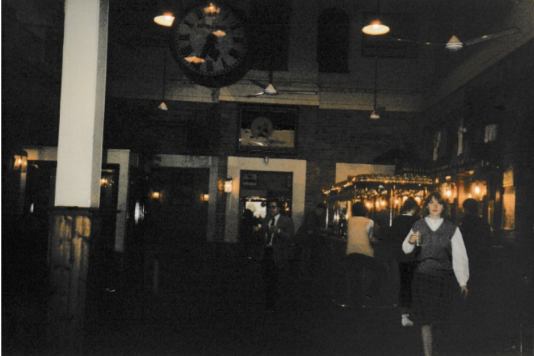 A grainy dark photograph taken inside a bar, a young woman standing on the right with a drink in her hand. There are warm lights behind her 