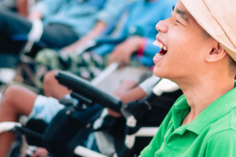 A young man is seated in a wheelchair and smiling. He is wearing a green polo shirt and a beige trilby hat