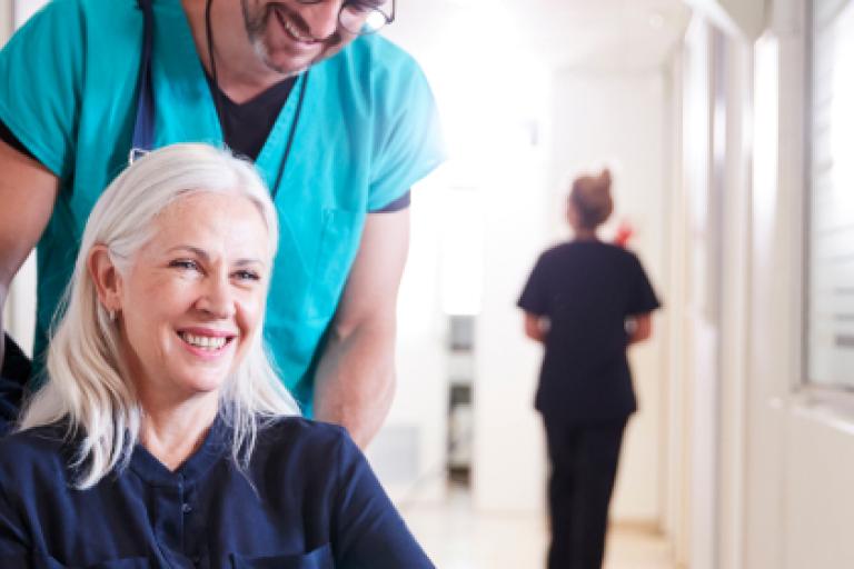 A woman with long white-blond hair is seated in a wheelchair. A man wearing medical scrubs is pushing the chair along a hospital corridor.