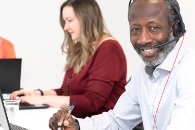 A man wearing a phone headset and working at a laptop is smiling at the camera. 2 women are working at laptops near him