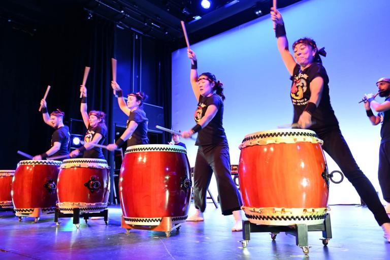 Four people wearing black stand in front of large red drums with their arms raised