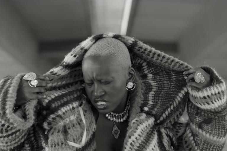 A black-and-white image of a person with cropped hair pulling up a patterned coat around their shoulders. They wear bold jewelry, including large rings, earrings, and layered necklaces, and their face shows a strong, emotional expression.