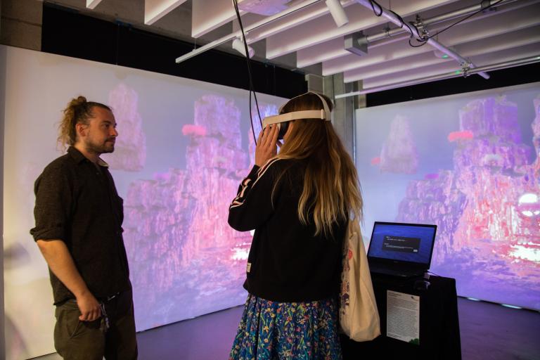 A woman wearing a VR headset interacts with a digital installation in a gallery space, while a man stands nearby observing. Behind them, large projected visuals show floating rocky landscapes in pink and purple tones with glowing elements. A laptop on a pedestal controls the projection.
