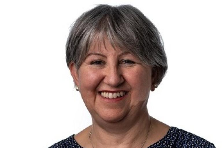 Angela is a woman with short grey hair. She is wearing a dark blue patterned top and a silver necklace