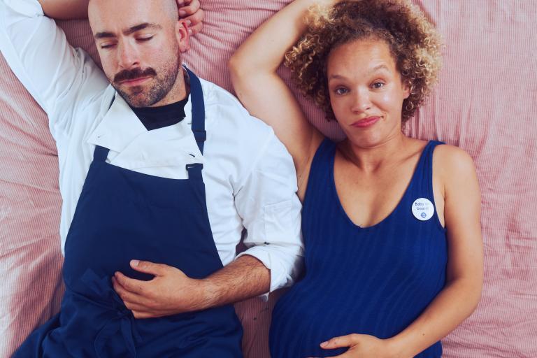 A man and woman lying on a bed, woman is looking straight to camera holding a baby bump.