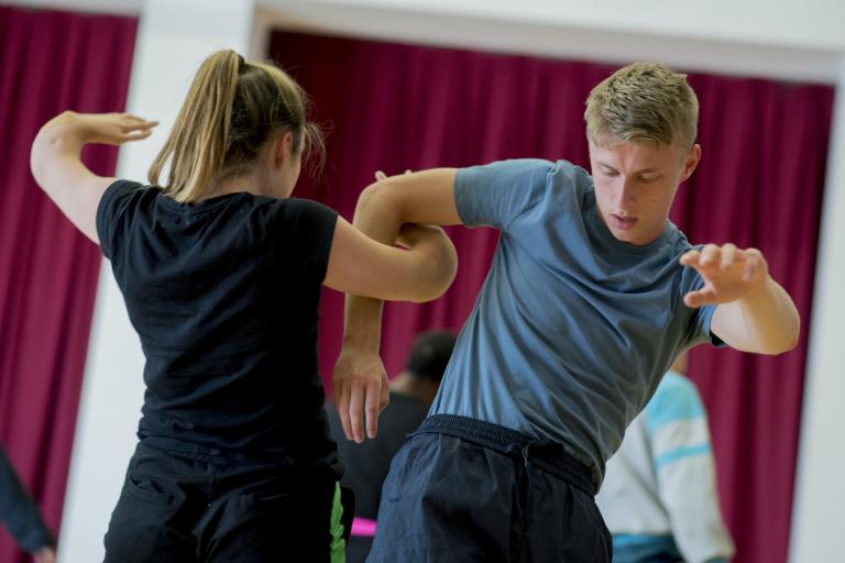 Two people dance closely together in an indoor workshop, their arms intertwined as one faces the camera and the other turns away.