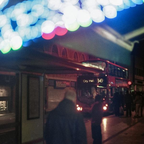 Rye Lane on a dark evening with the 343 bus passing closed shop fronts. A shop sign made up of LED lights twinkles in the foreground