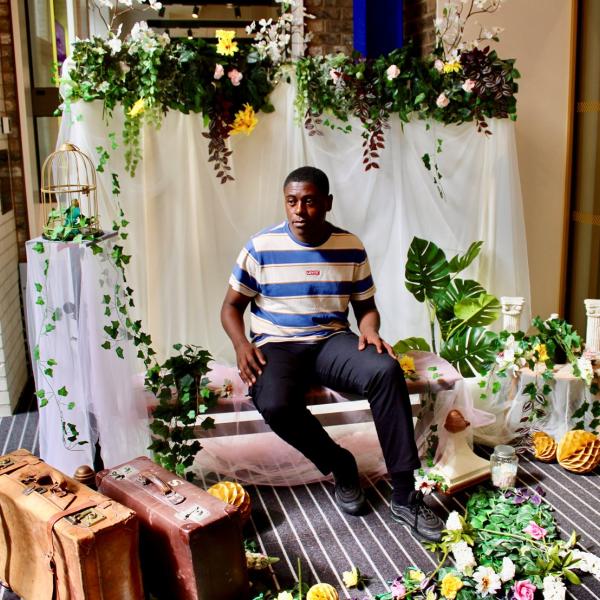 A man sitting in a photo studio with colourful potted plants, a gold bird cage, classical columns and a draped backdrop