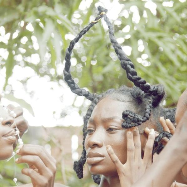 Close-up of the faces and hands of three young Black women with intricately braided and styled hair. They are outside among trees.