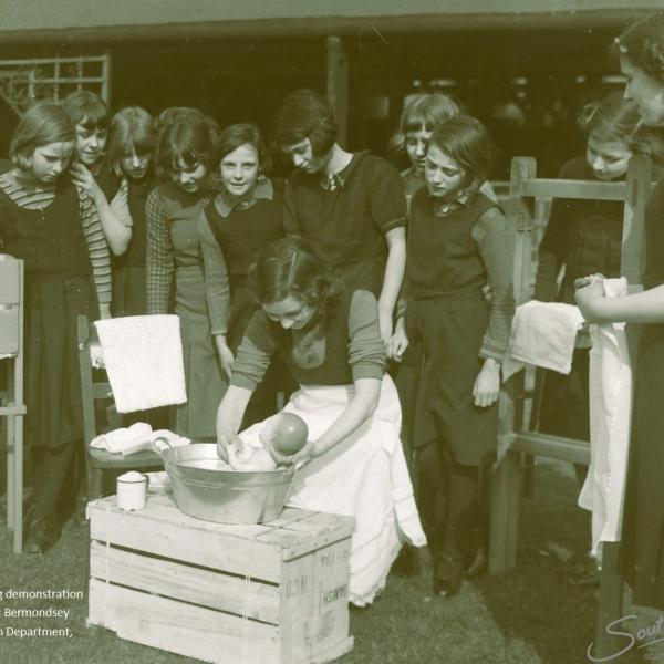 Black and white image showing group of young girls watching a demonstration of how to bath a baby.