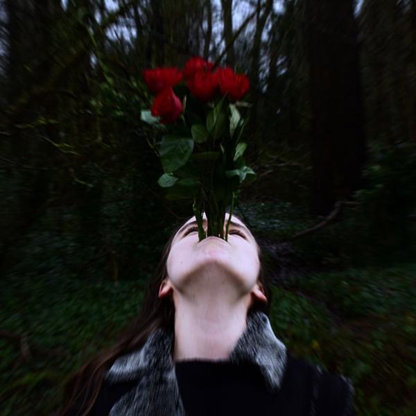 A young woman, looking up holding a bunch of roses by the stems with her mouth, against a woodland backdrop. 