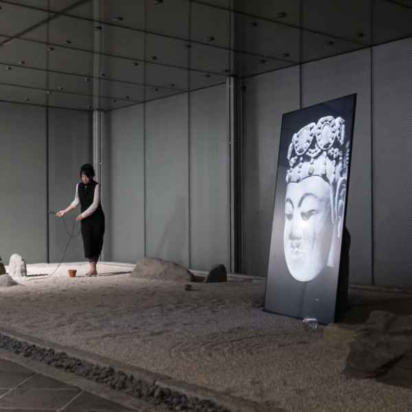 An artist performs in an art installation. They walk on sand, and there is a screen.
