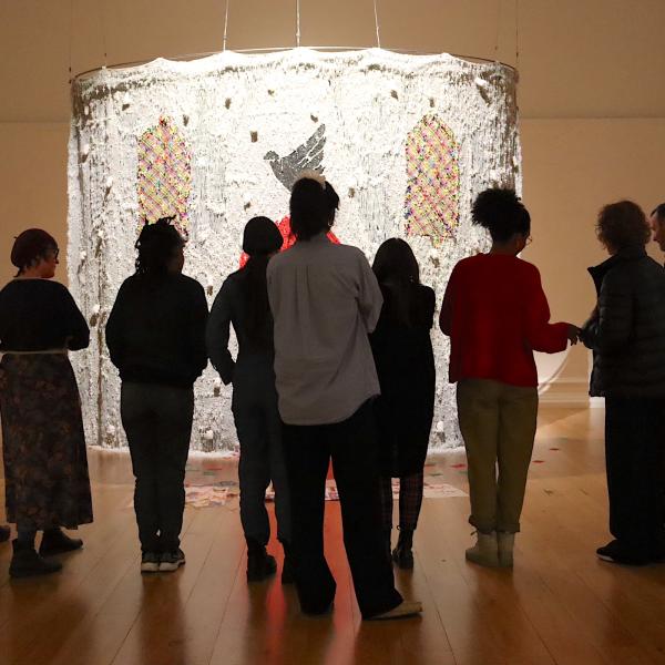 People stand in a dark art gallery. They are gathered around a large installation that is spot lit. The installation is an alter-type structure made of beads. 