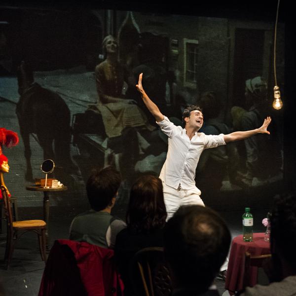 A theatrical performer in white gestures expressively on stage in front of a seated audience.