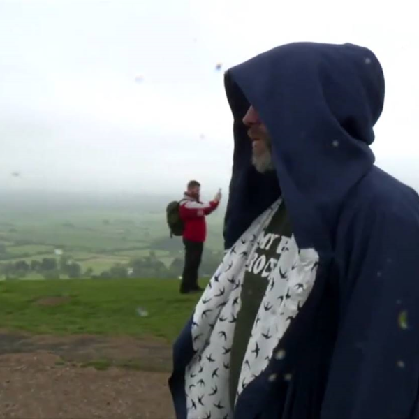 An image of Wes Viola in a dark blue hooded robe. In the back ground are the fields of Somerset, and a man wearing orange taking a picture, who due to an illusion of perspective appears much smaller than the figure of Wes in the foreground