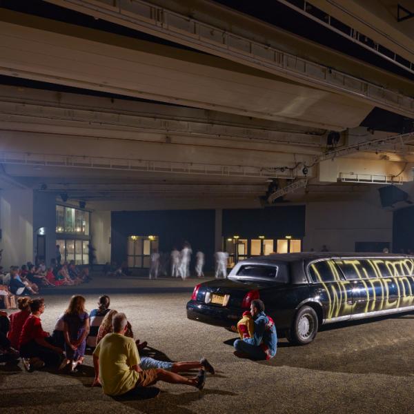 Audiences sit in front of a black limousine in a concrete space.