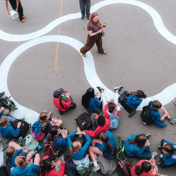 A birdseye view of a group of schoolchildren sitting down on the floor being spoken to by a facilitator who is standing in front of them