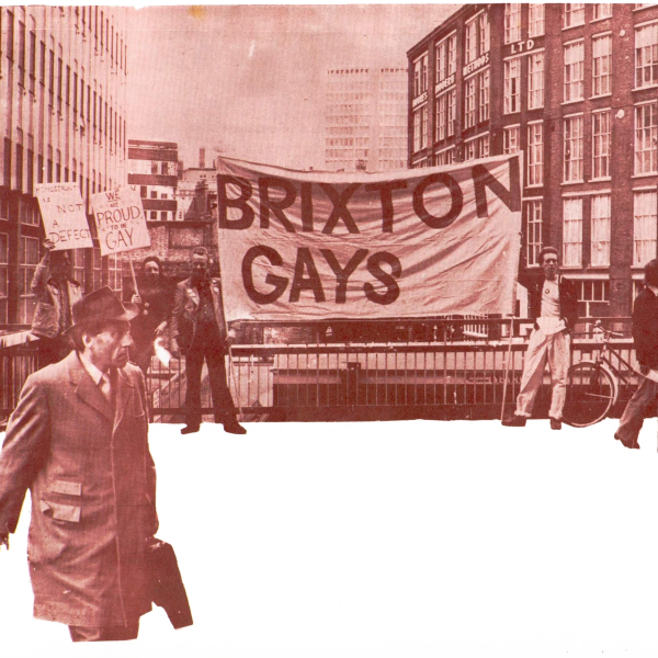Protesters hold a large "Brixton Gays" banner and signs reading "We are proud to be gay" and "Homosexuality is not a defect" on a city street, while a man in a coat walks past.