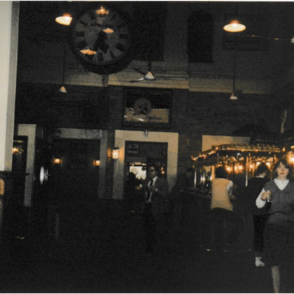 A grainy dark photograph taken inside a bar, a young woman standing on the right with a drink in her hand. There are warm lights behind her 