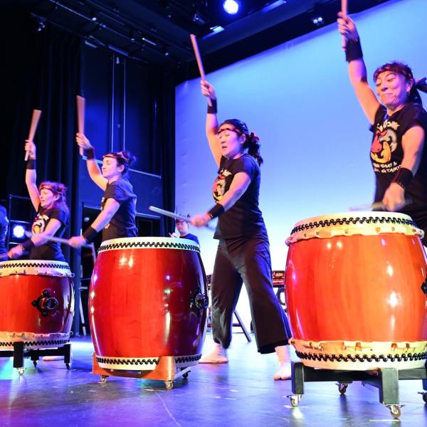 Four people wearing black stand in front of large red drums with their arms raised