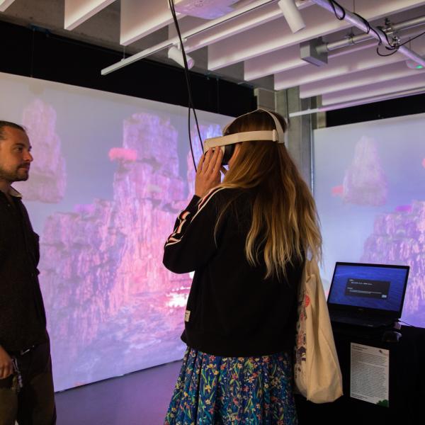 A woman wearing a VR headset interacts with a digital installation in a gallery space, while a man stands nearby observing. Behind them, large projected visuals show floating rocky landscapes in pink and purple tones with glowing elements. A laptop on a pedestal controls the projection.