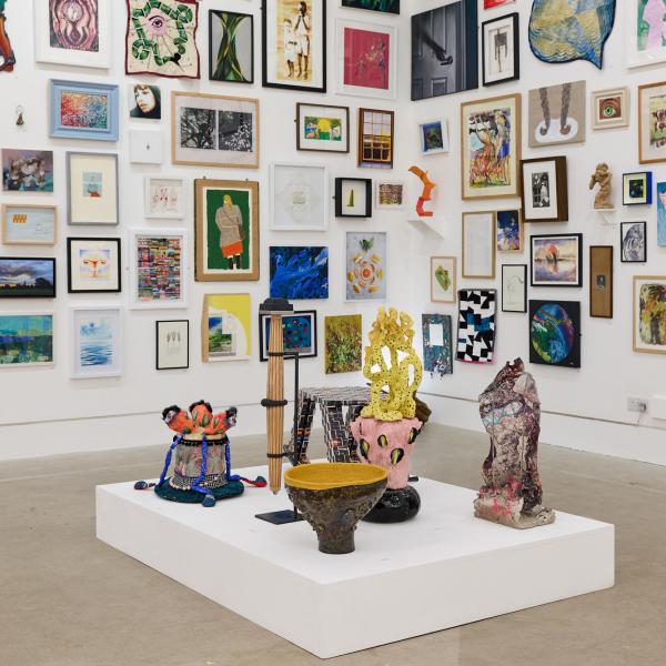 eclectic salon style hanging artworks on white walls, a white plinth with an assortment of sculptures in the foreground.