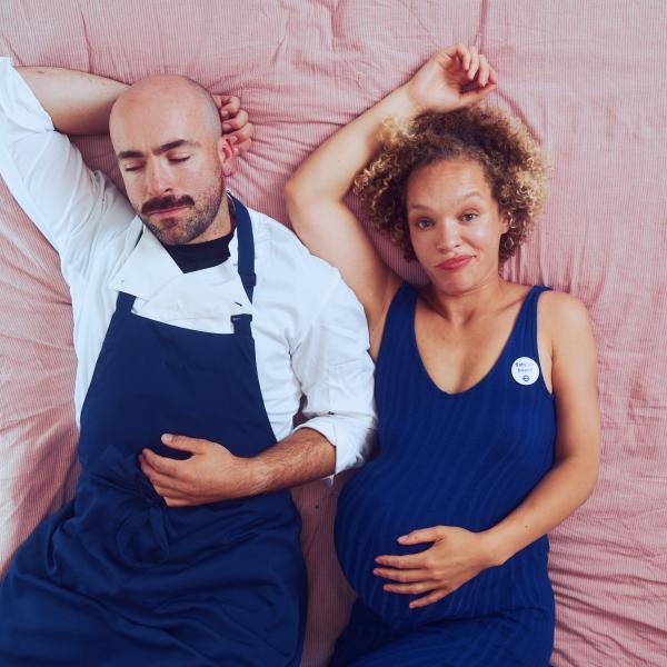 A man and woman lying on a bed, woman is looking straight to camera holding a baby bump.