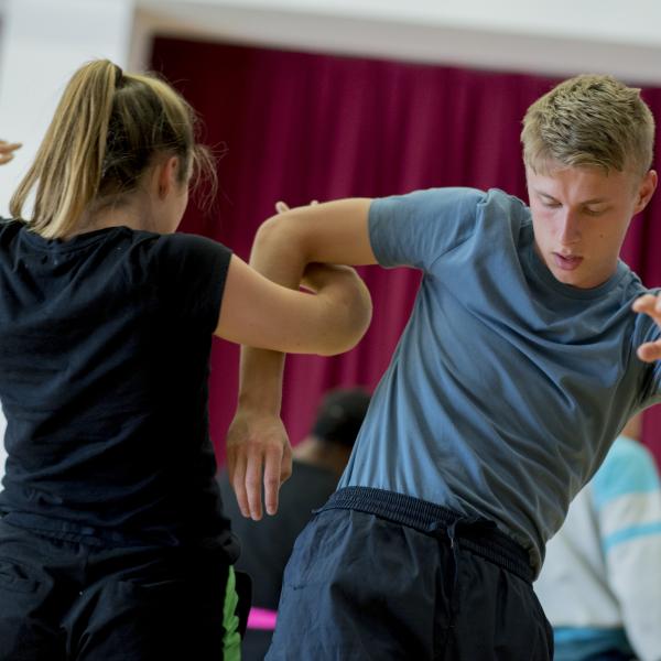 Two people dance closely together in an indoor workshop, their arms intertwined as one faces the camera and the other turns away.